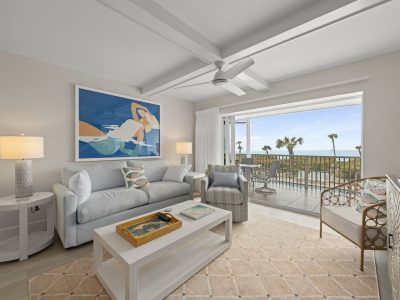 Bright coastal living room with a light gray sofa, abstract blue artwork, and open balcony doors to an ocean view.